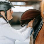 Adult woman prepares horse for riding by adjusting saddle in stable.